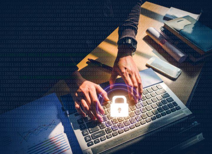 Cybersecurity professional working on a laptop at a desk with a large yellow lock illuminated and superimposed over it. Only the worker's hands are visible poking through a shadow.