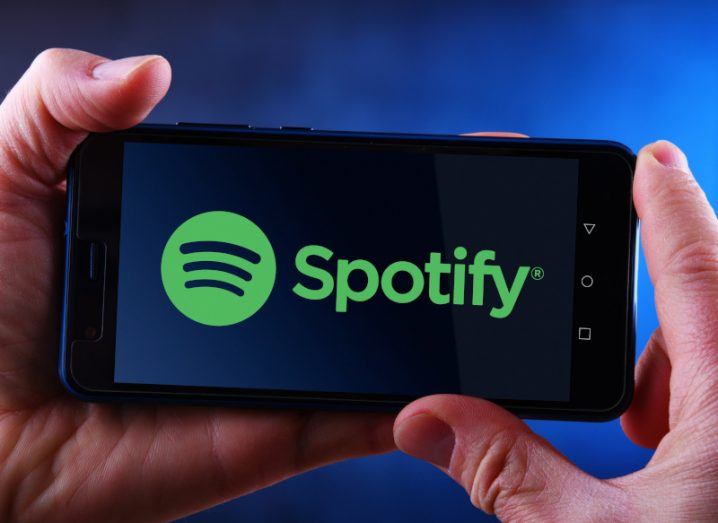 Spotify logo on a mobile phone held by two hands with a blue background.