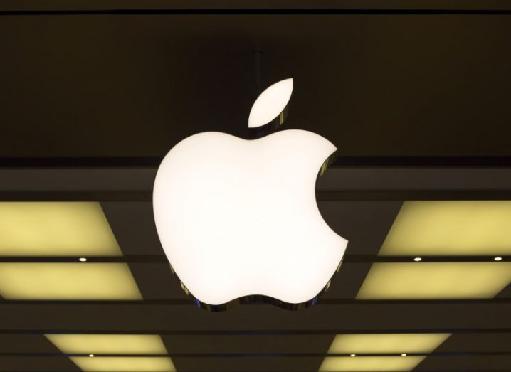 Apple logo lit up in front of a building, with some ceiling lights in the background.