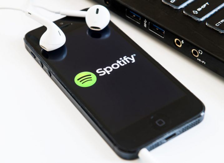 Smartphone with the Spotify logo displayed lying on a white table with white earphones.