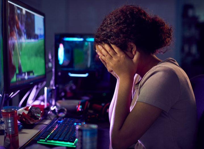 A young woman sits at a computer monitor with a video game on screen. She is visibly upset with her head in her hands.