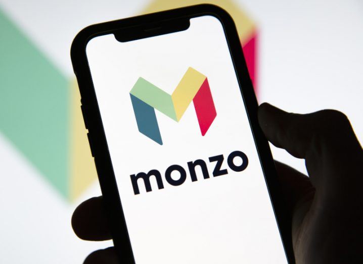 Monzo logo in a white background on a smartphone being held by a hand, with the logo also in the background of the image.