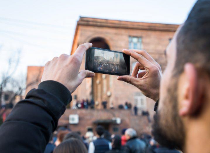 Over-the-shoulder view as a man films a protest taking place using his smartphone.