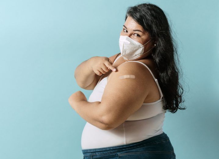 A woman poses like Rosie the Riveter with her bicep flexed. She is wearing a mask and has a plaster on her arm, indicating that she has just received a Covid-19 vaccine.