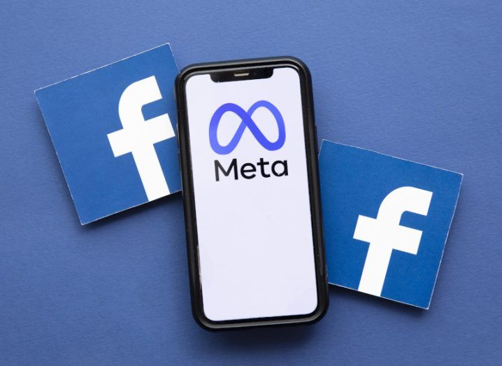 Smartphone displaying Meta logo flanked by two Facebook logo cards against a blue background.