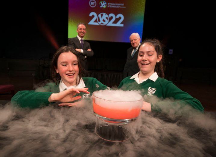 Two girls crouch behind a table with a bubble of smoke emerging from a science project.