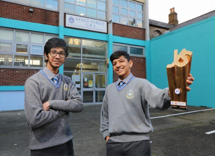 Two teenage boys stand in front of Synge Street school, with one of them holding a gold BT Young Scientist trophy.