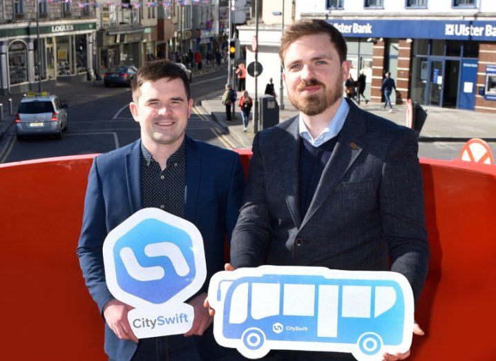 Two men standing on the rooftop of a red bus with CitySwift signs smiling.