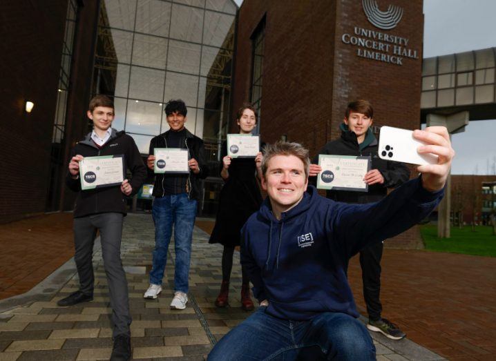 John Collison stands crouches in front of the University Concert Hall Limerick to take a picture of himself and four teenagers standing behind him, all holding up TECS certificates.