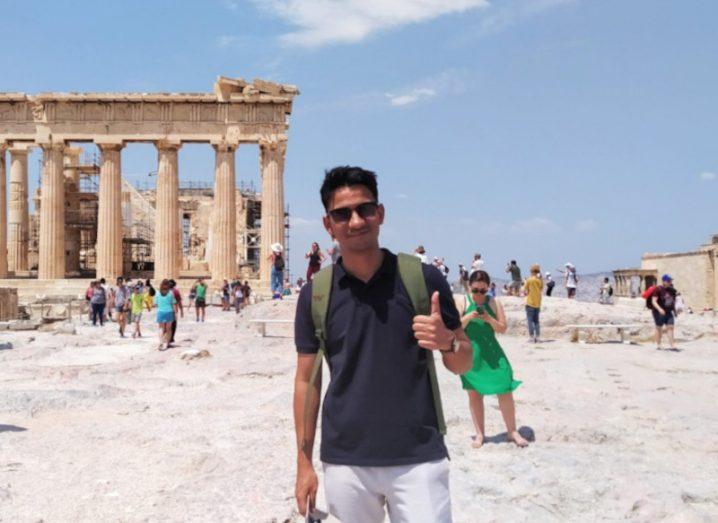 A man stands in summer clothes in front of an ancient ruin.