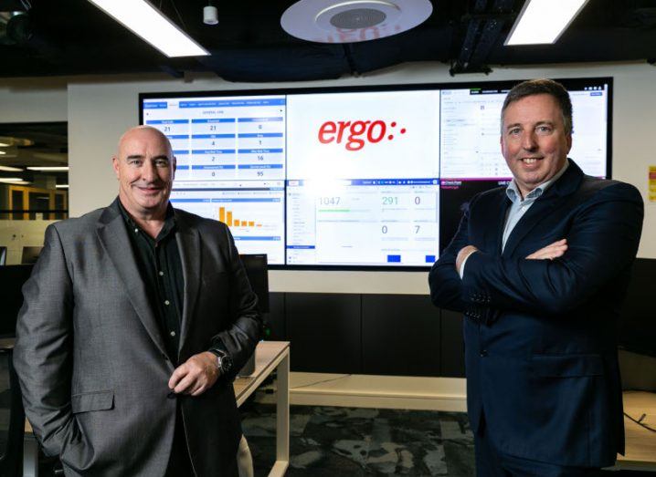 Two men stand in front of a wall of screens featuring the Ergo logo.