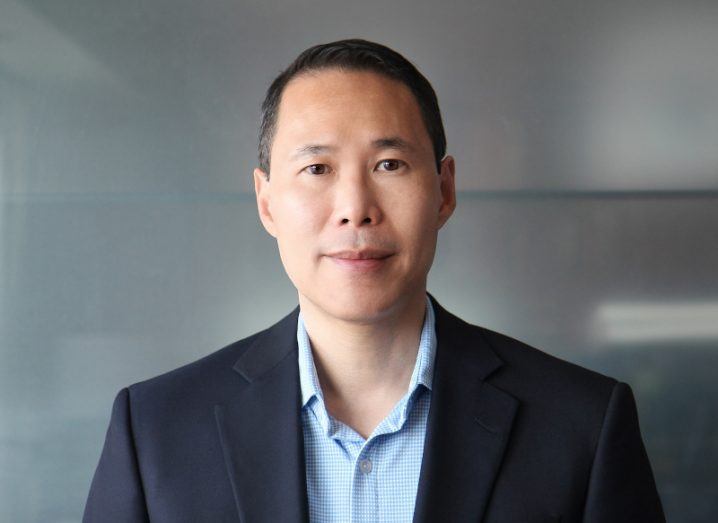 A man in a dark suit jacket and a light blue shirt smiles at the camera.