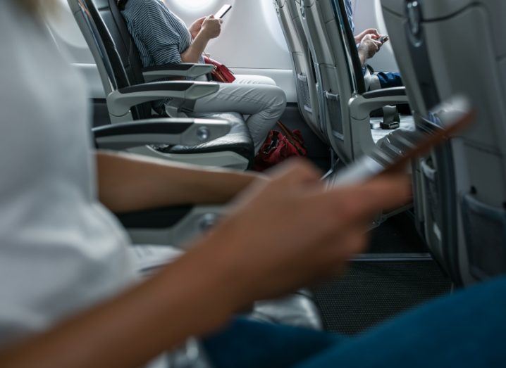 Person inside a plane holding their smartphone in hand. Another person in the background does the same.