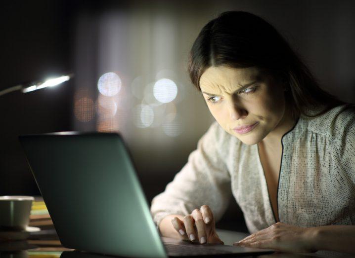 Woman looks suspiciously at a laptop screen in a dark room with a table light switched on.