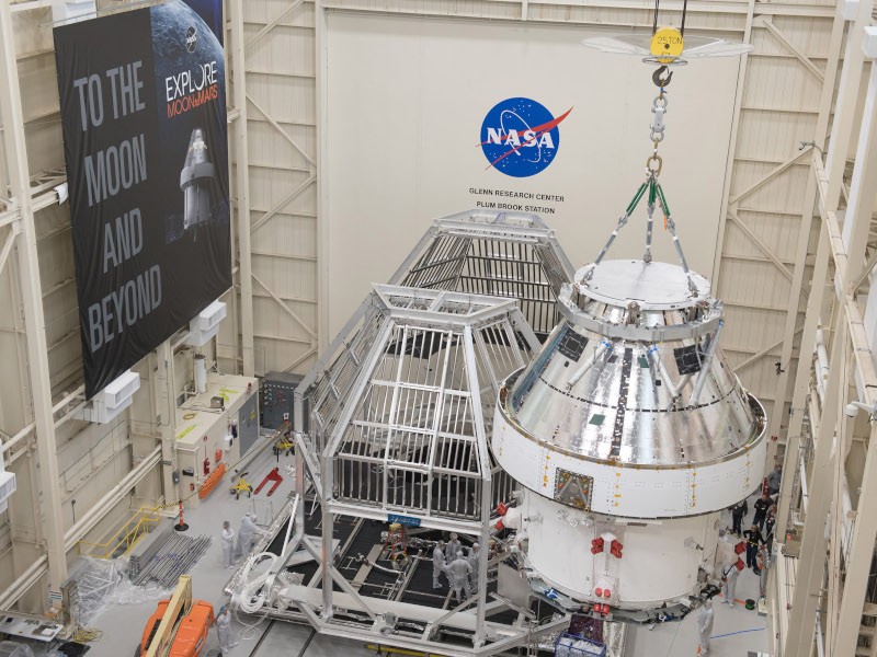 Part of a spacecraft is lifted into a testing chamber in a large room, with the NASA logo and a poster about moon exploration in the background.
