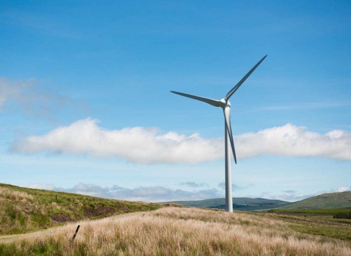 A wind turbine on hilly and grassy land with blue sky in the background.