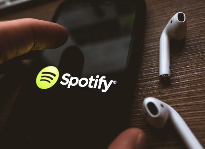 Spotify logo on a smartphone screen with a finger touching the screen. Wireless earphones lay to the right of the phone.