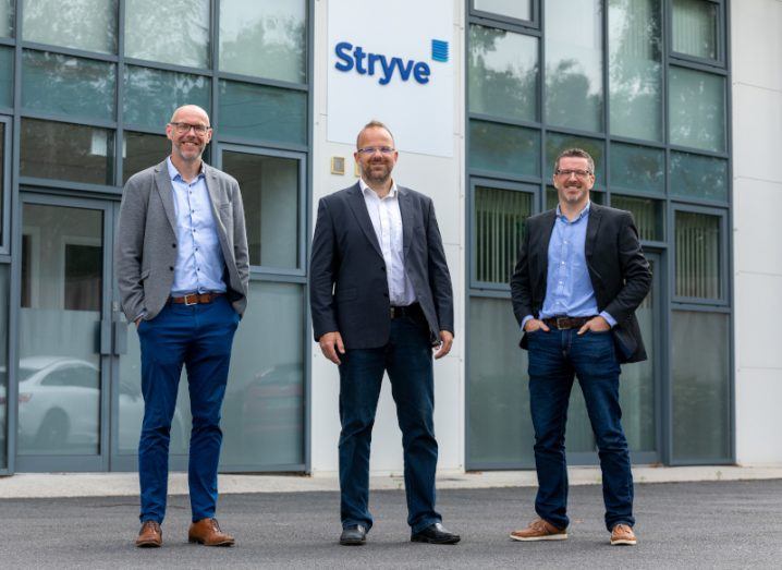 Three men stand side-by-side in front of a building with the Stryve logo on it.