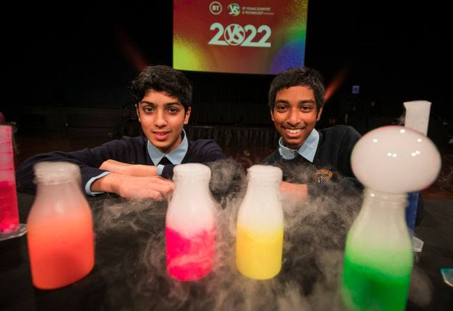 Two boys in school uniform crouching behind a table with multi-coloured chemicals in lab flasks with gas bubbles coming out.