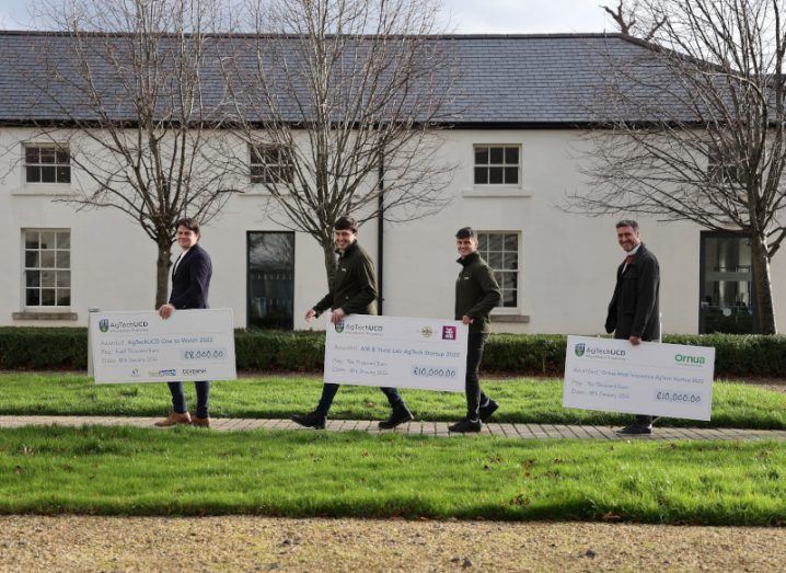 Four men holding big cheques in their hands walk across a path on the UCD campus.