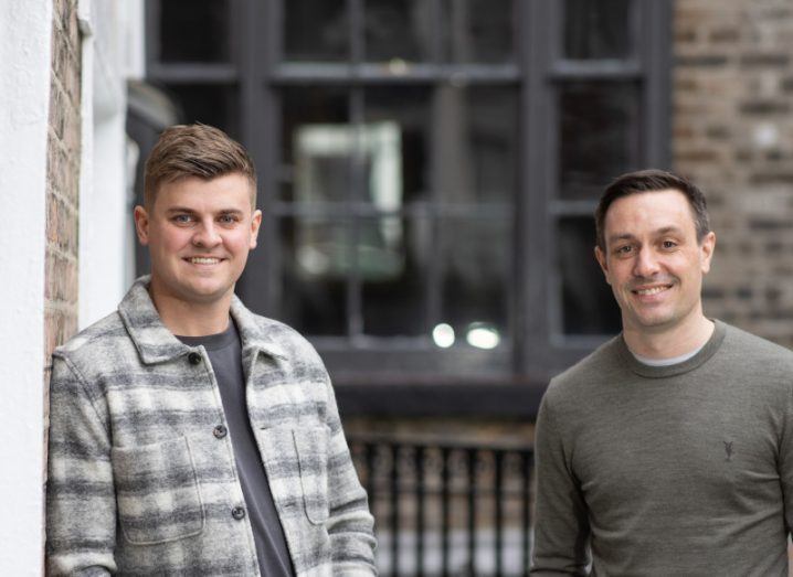 Two young men pose for the camera with blurred windows in the background.