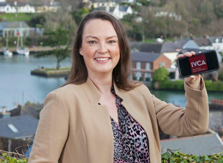 Director general of the Irish Venture Capital Association Sarah Jane Larkin holding a phone with the company's logo on it, with a river in the background.