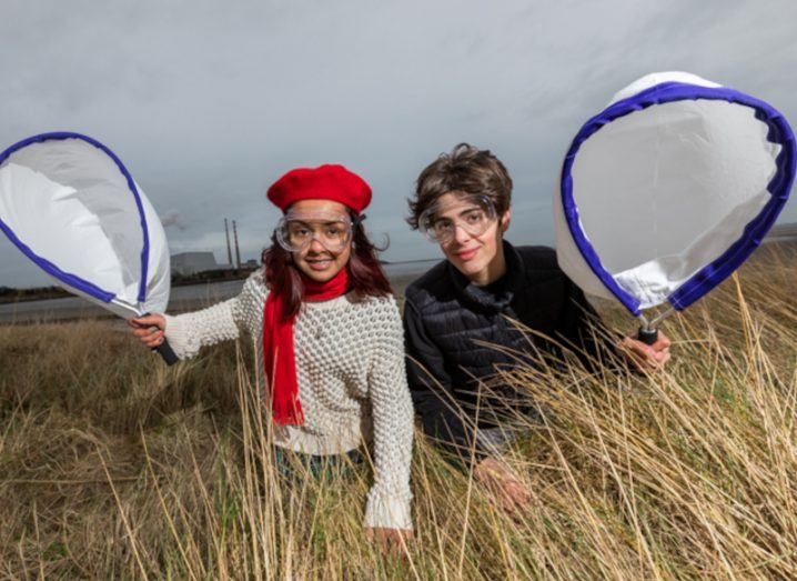 Two teenagers crouching in grass holding big circular nets in the air and looking at the camera.