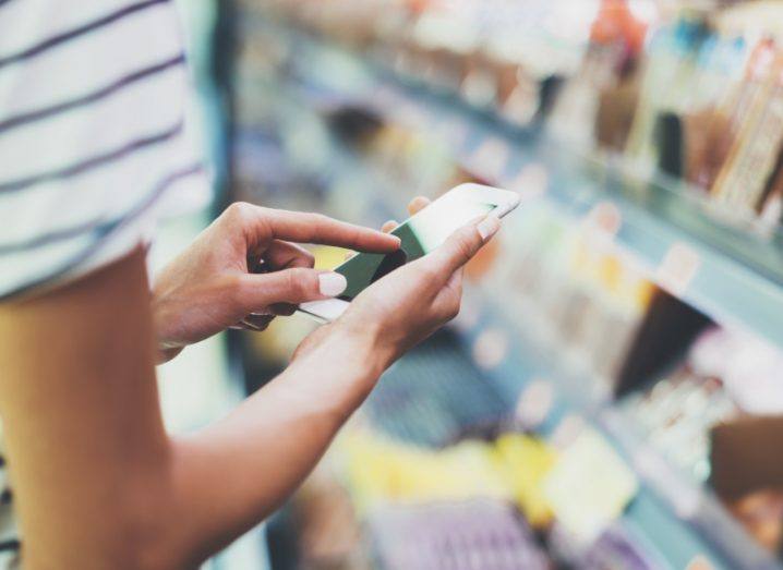 Person shopping on a mobile phone in their hands, with shop isles in the background.