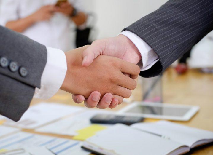 Two business people shaking hands with a table covered in books in the background.