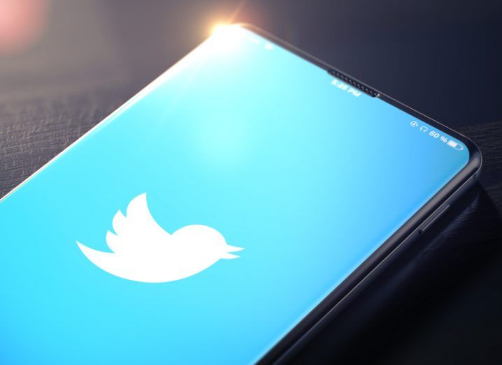 Twitter logo with a blue background on a mobile phone screen, resting on a dark table with some sunlight reflecting off the corner of the phone.