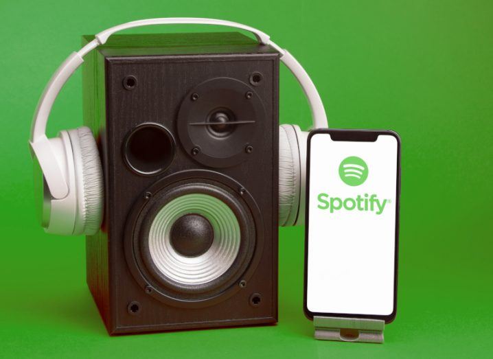 Spotify logo with a white background on a mobile phone screen, standing next to a music speaker with headphones on it.