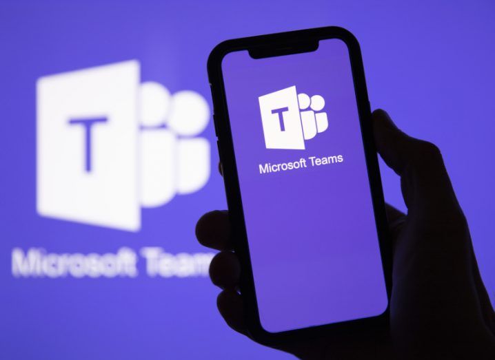 Person's hand holding a phone with Microsoft Teams logo displayed on the screen and in the background.