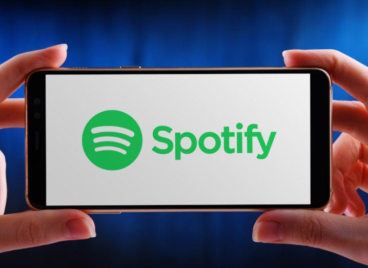 Spotify logo on a white background on the screen of a mobile phone being held by two hands.