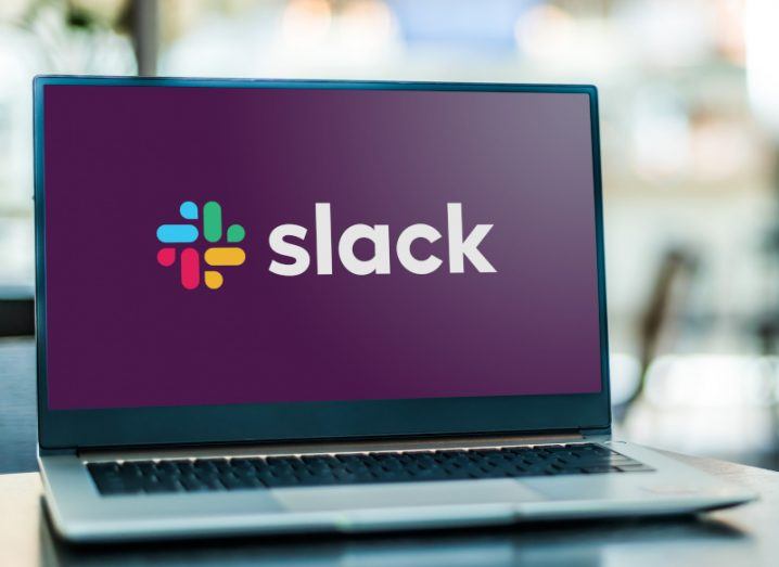 The Slack logo is displayed on a laptop placed on a desk with an office out of focus in the background.