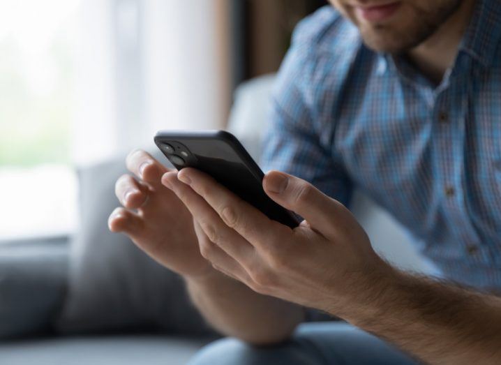 Man sending a text on a mobile phone with a window in the background.