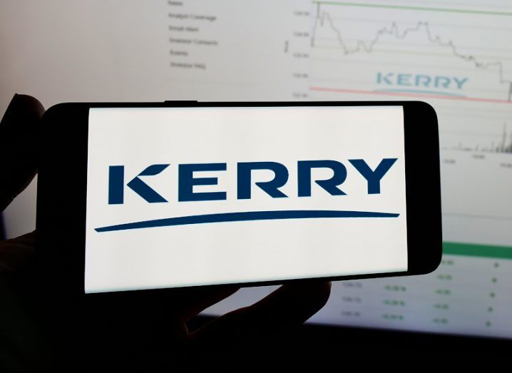 Kerry Group logo on a white background of a mobile screen being held by a person with a screen on in the background.