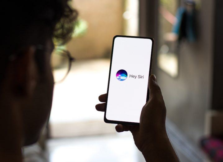 Siri logo with the words "Hey Siri" next to it on a mobile phone screen being held by a person.