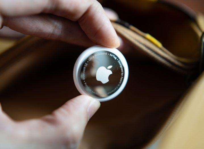 An Apple AirTag being held in between a finger and a thumb with a wallet in the background.