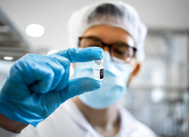 Researcher holding a lab made vaccine vial in his hand, while wearing blue gloves and a face mask.