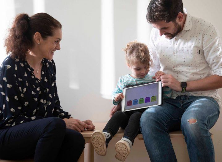 A woman sits on a bench observing a small child using a tablet app with the assistance of their father.