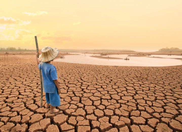 Child wearing a hat standing on dry, cracked ground staring at shallow water in the distance.