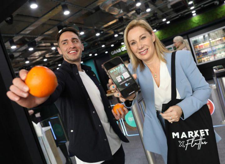 Greg O'Shea holding an orange at the camera while Josepha Madigan stands next to him holding a smartphone with "Market x Flutter" written on it.