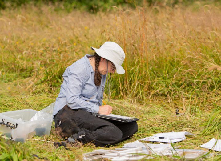 A woman sitting on the grass in the middle of a field surrounded by research papers. She is writing on a clipboard.