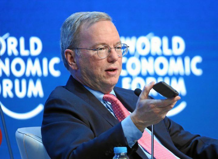 SandboxAQ investor Eric Schmidt speaks on-stage in front of a blue World Economic Forum backdrop, gesturing with his phone is his hand.