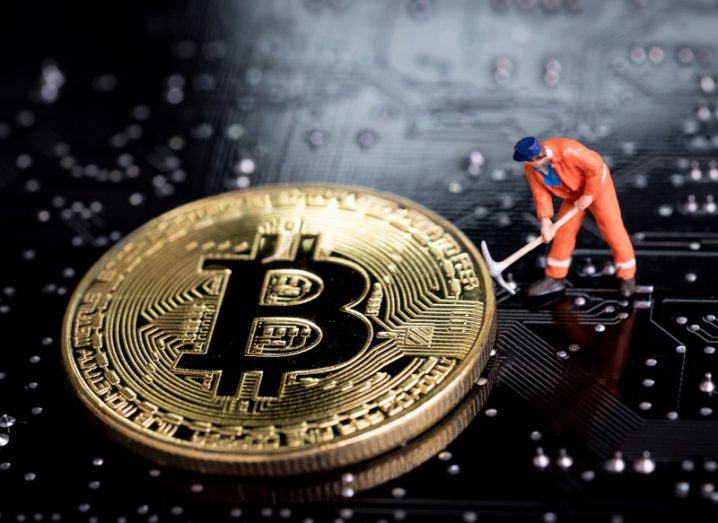 A miniature model of a person mining with a pickaxe on the surface of a computer motherboard. The pickaxe is touching a golden coin with the bitcoin icon on it.