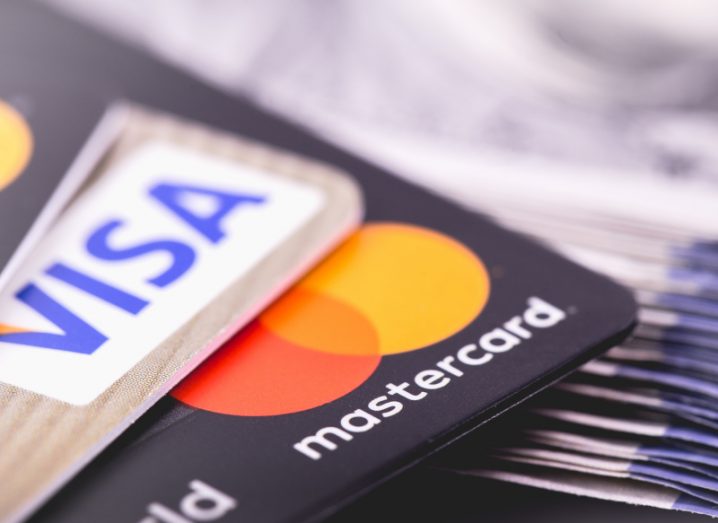 A Mastercard card and a Visa card are sitting on a pile of dollar bills.