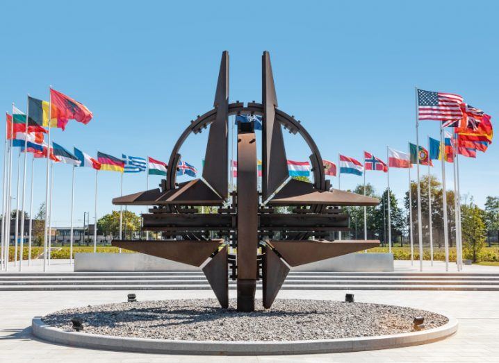 NATO monument in Brussels with multiple flags behind it and a clear blue sky overhead.