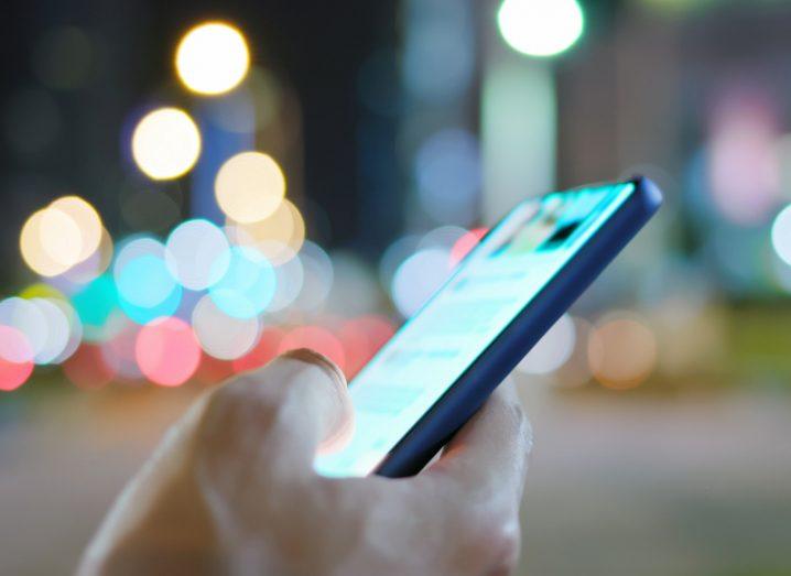 A person is using a mobile phone on a city street with blurred lights in the background.