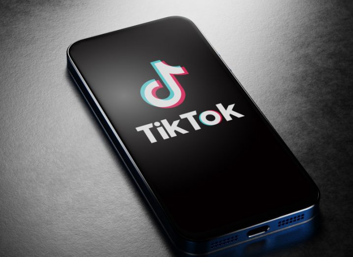 TikTok logo on a black background on a mobile phone screen, laying on a dark surface with some light shining on it.