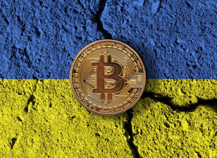 A gold bitcoin against a broken wall with the Ukrainian flag painted on it.